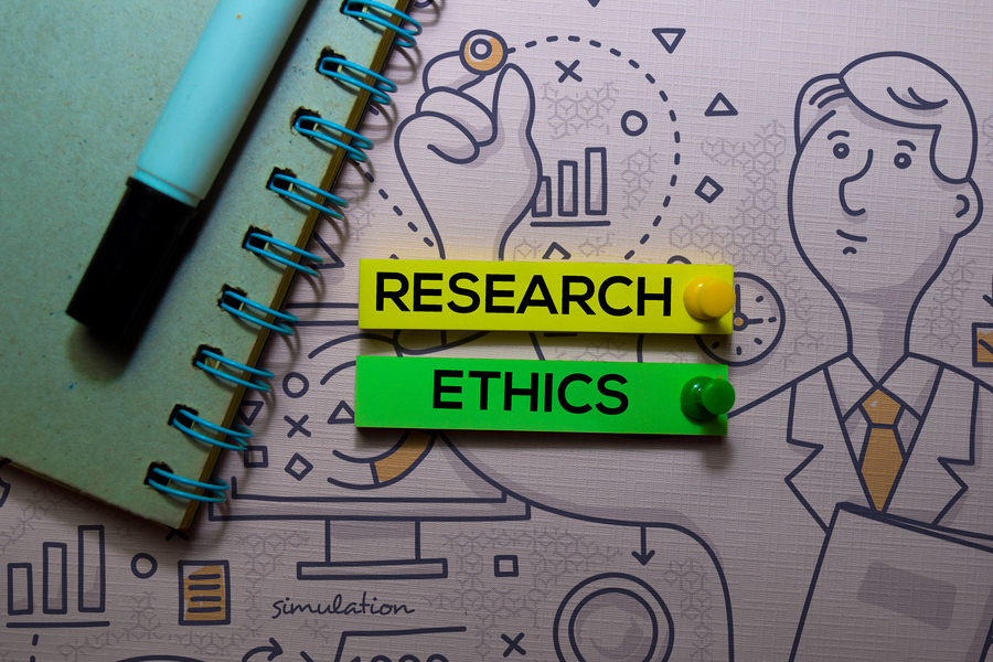 Photo of an illustration of a man wearing a lab coat with the words “Research” and “Ethics” pinned to it. A notebook and pen appear on top of the illustration.