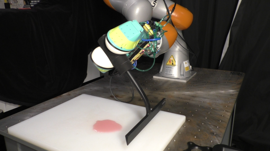 Photo of a robotic arm holding a squeegee in preparation to wipe up a blob of pink liquid