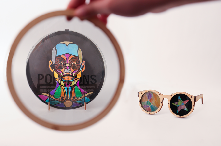 At right are wooden glasses with colorful stained-glass-style mosaic lenses. At left, a hand holds a wooden circle, inside of which appears a stained-glass style mosaic image of a human face and shoulders, with the word POLAGONS superimposed