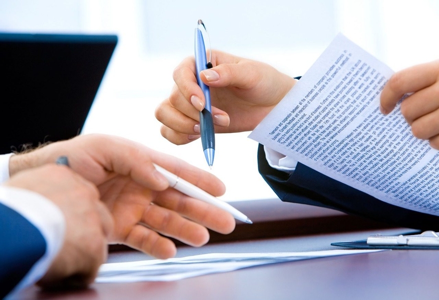 A closeup photo of two sets of hands. Both hold pens, and one holds a piece of paper.